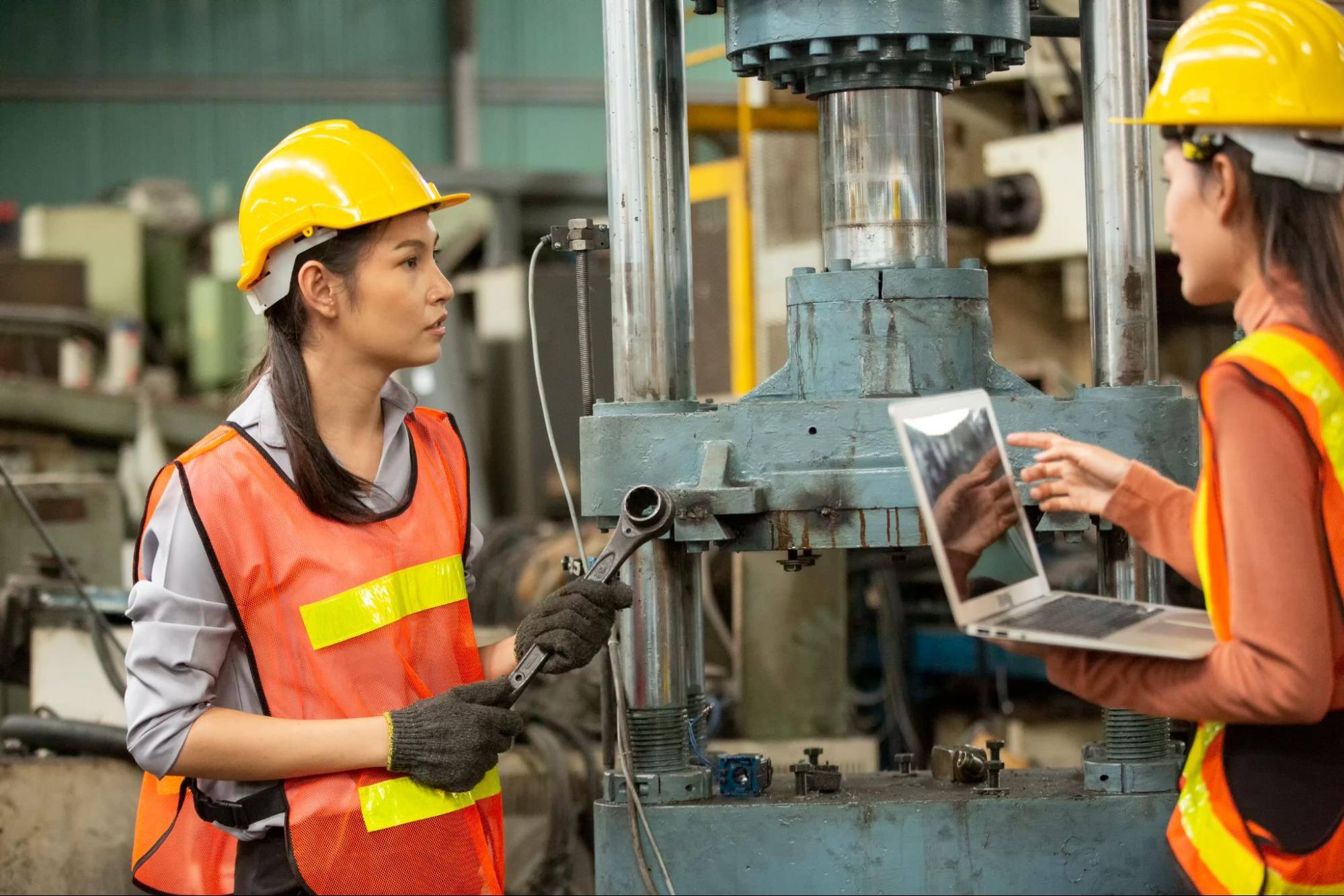 Two female construction workers with one holding a tool and the other holding a laptop.&nbsp;