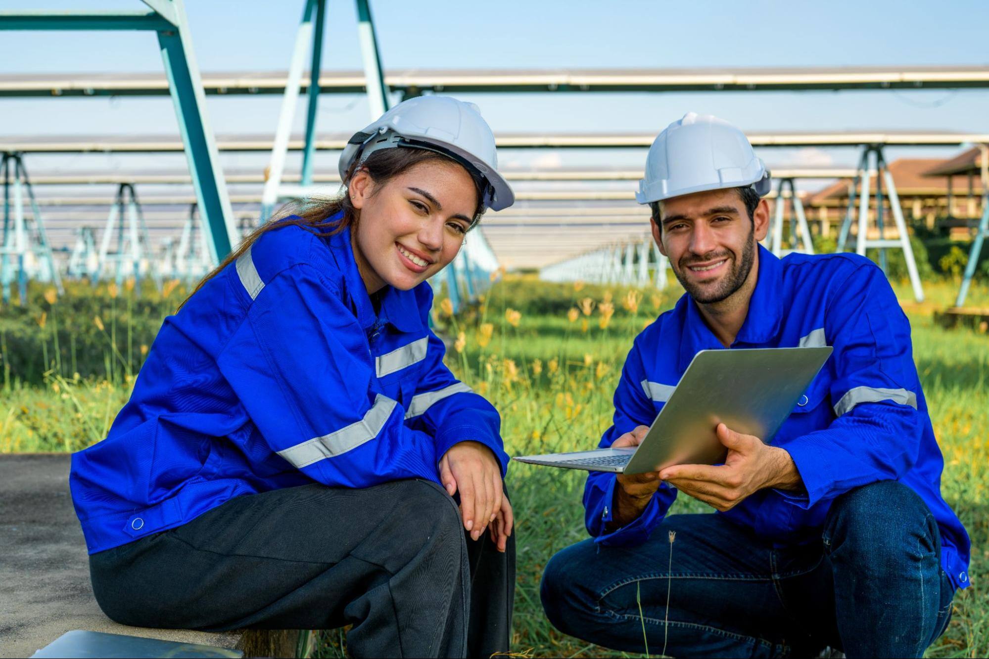 A man and a woman in a field with a laptop both wearing blue uniforms and hardhats looking toward the camera and smiling.