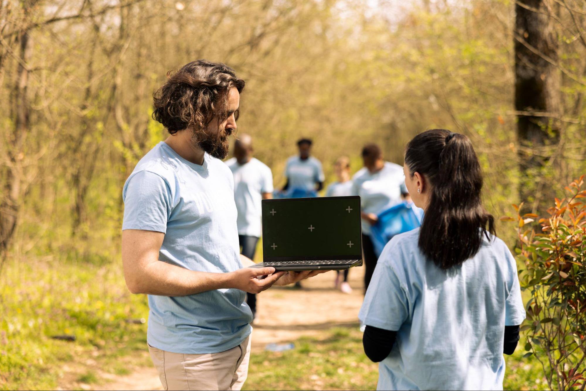 A man and woman face each other while the man holds an open laptop in the field with other workers in the background.