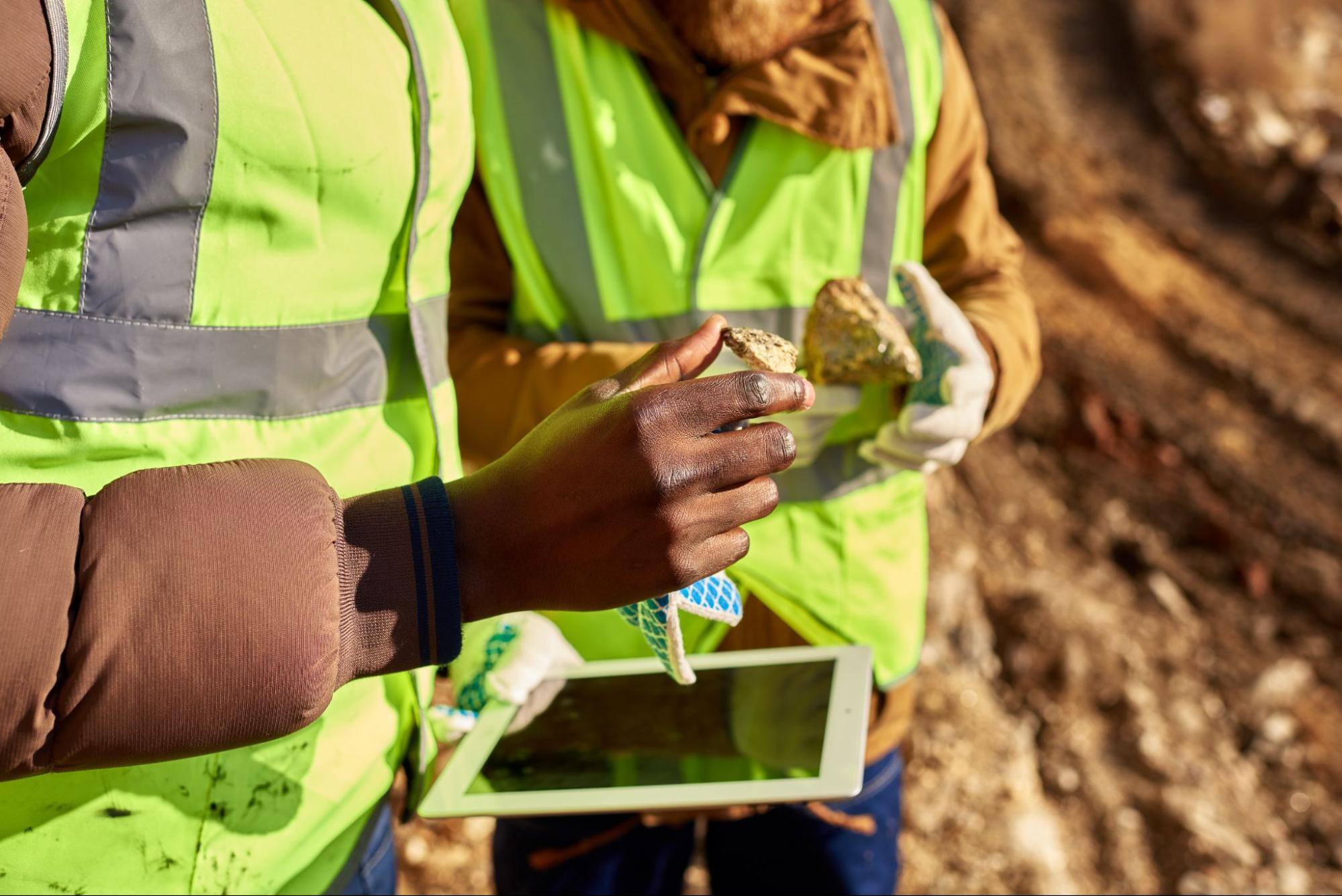 A cropped view of two men's hands holding rocks and one of them having an intelligent tablet in the field.