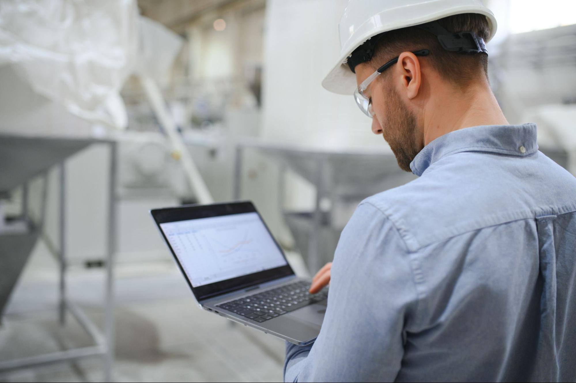 A man wearing a white hard hat and safety goggles standing with his back to the camera and working on a laptop.