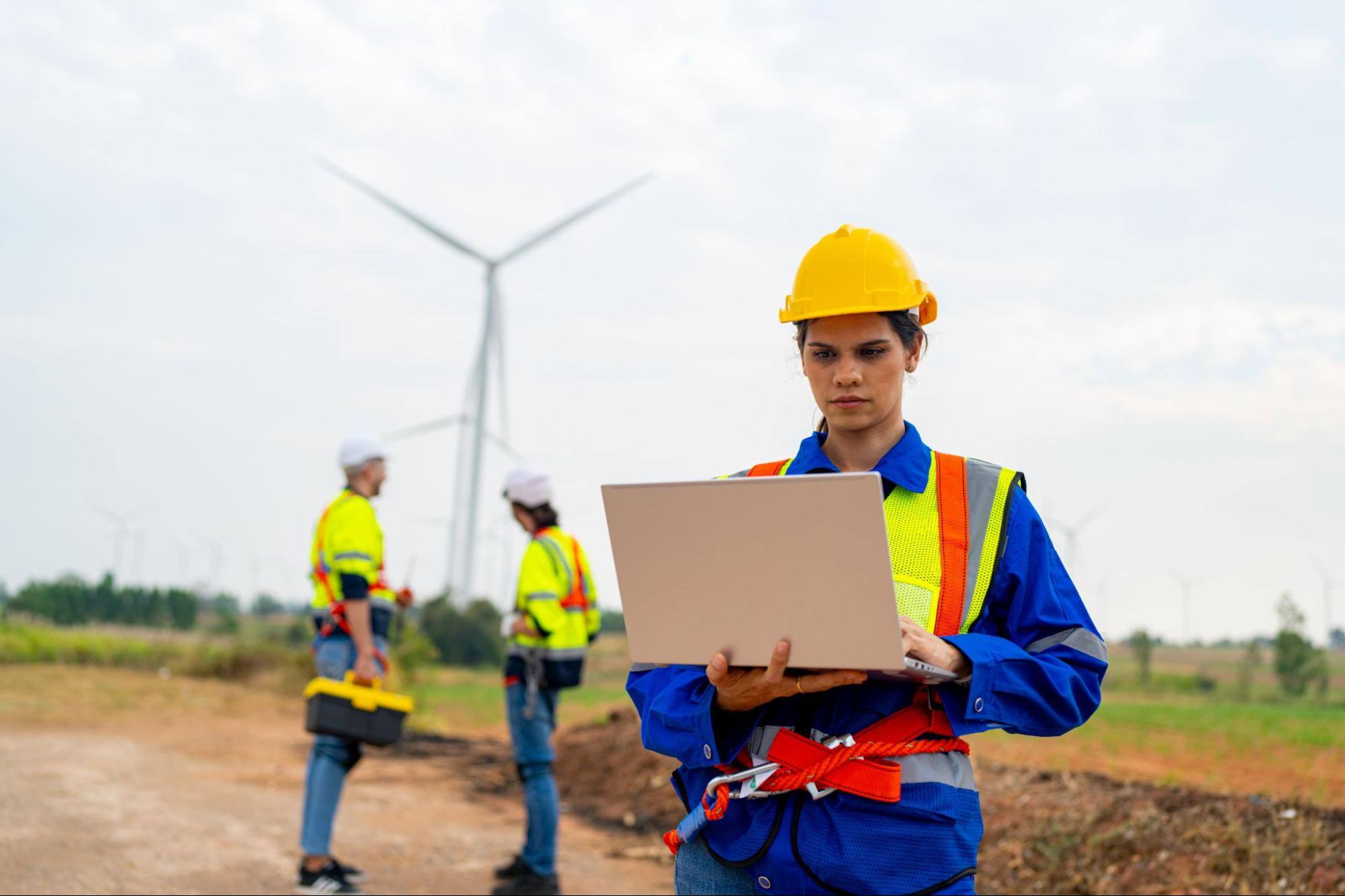 Woman in a hard hat looking at a computer next to two men and a wind turbine.