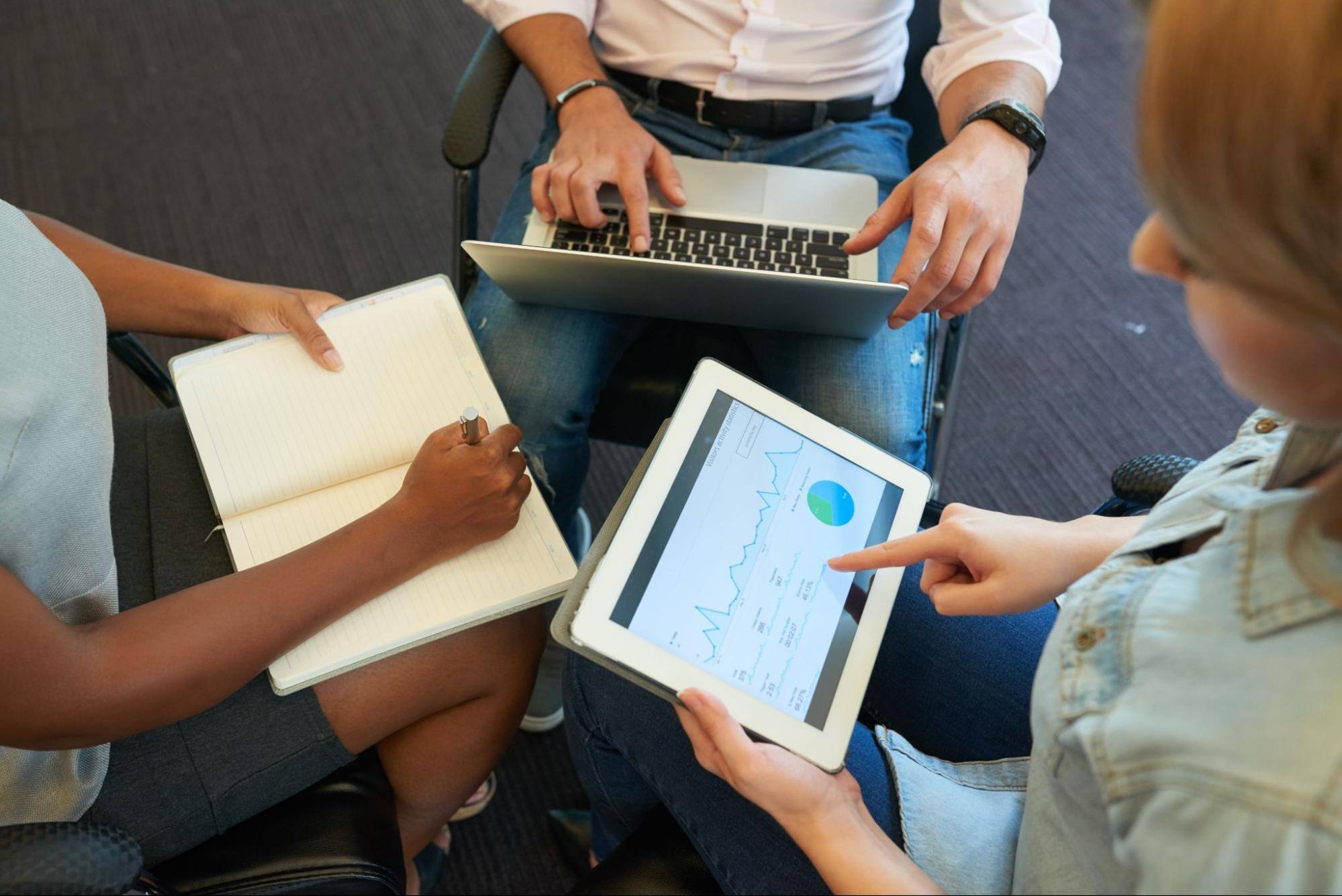 View from above of three women using different mediums for data collection–a notebook, a smart tablet, and a laptop.