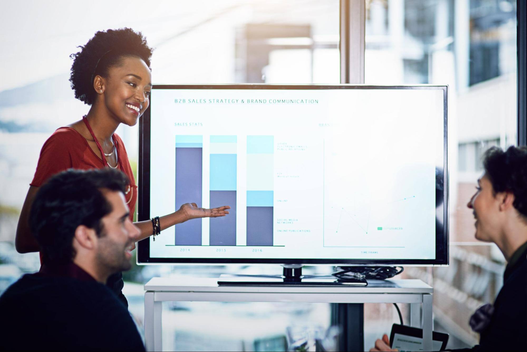 A woman is standing up and giving a presentation with data on a computer screen while two men are sitting down and looking at the screen.