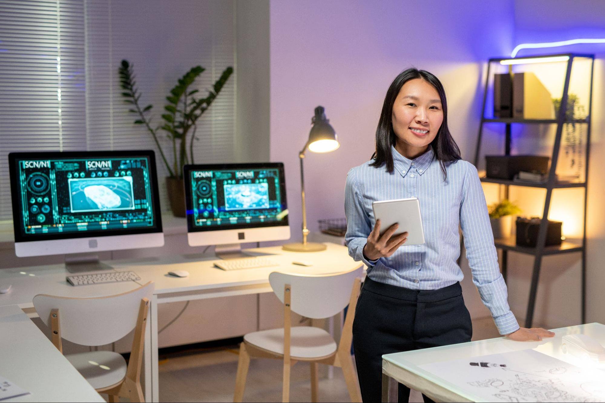An Asian woman holds a tablet while presenting software to improve resource management for a company. Two computers on a white desk are visible behind her in a modern, stylish office.
