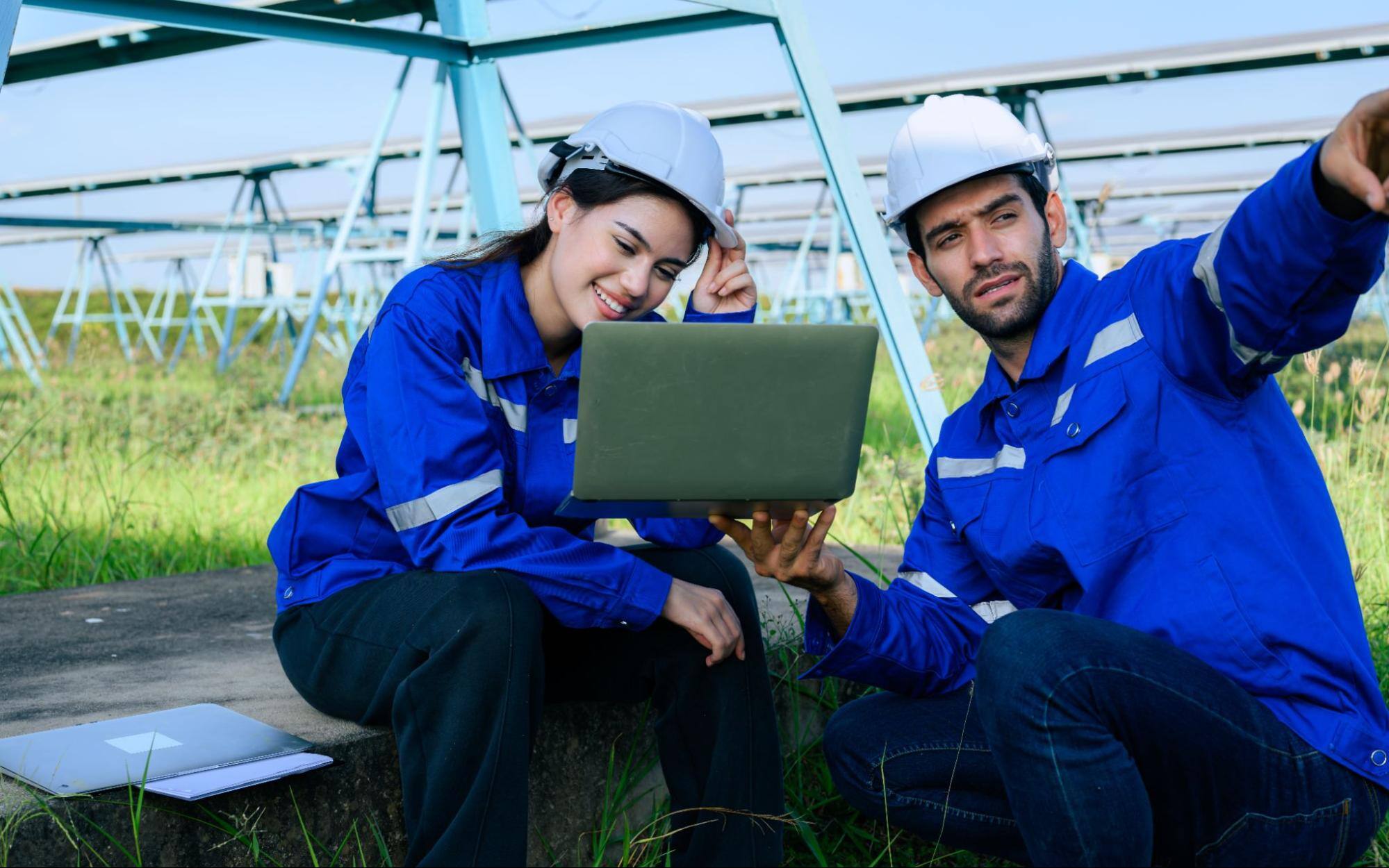 Two workers in hard hats looking at a computer next to solar panels. 
