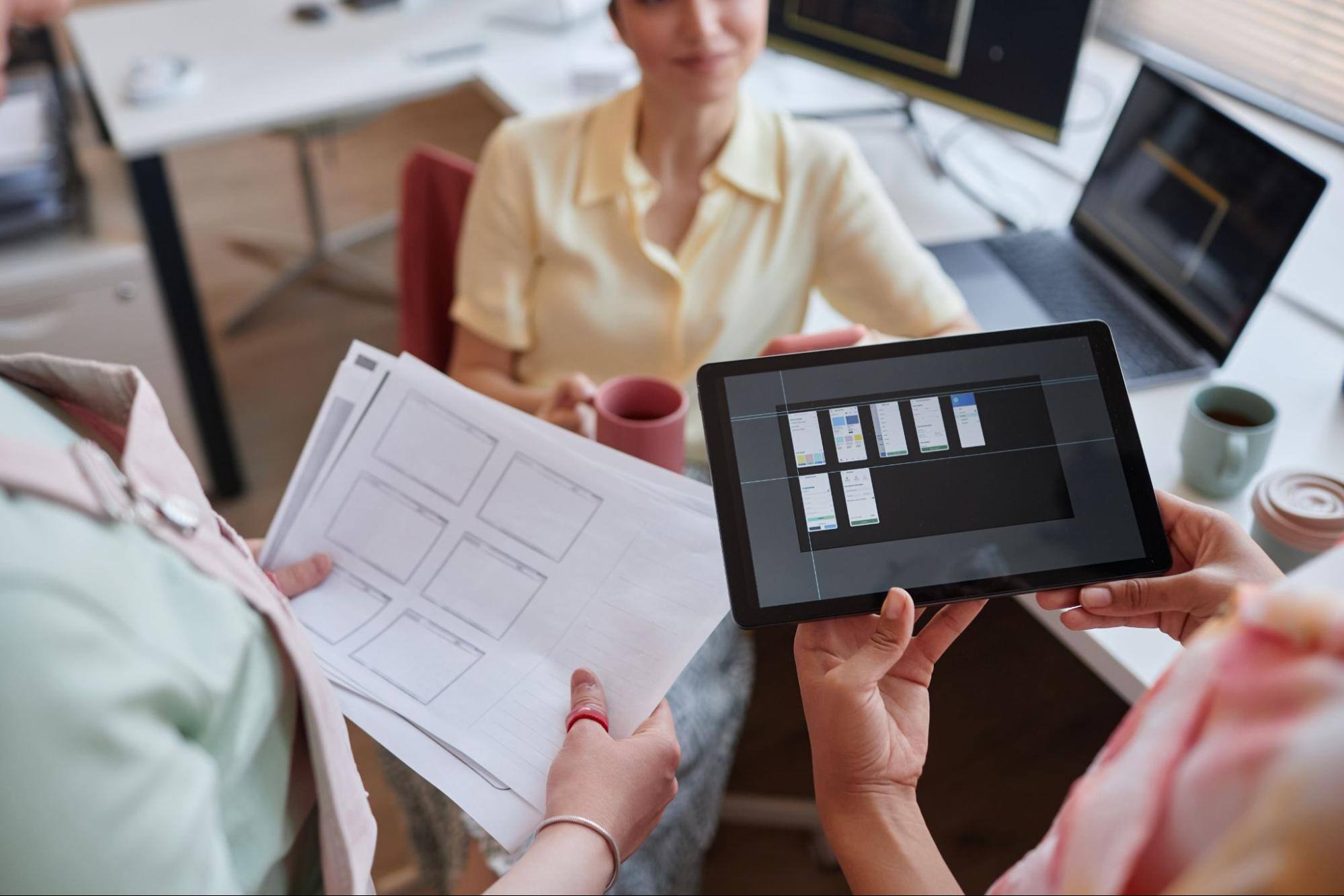 Two young women review project management software features on a digital tablet while sharing sketches with their boss.