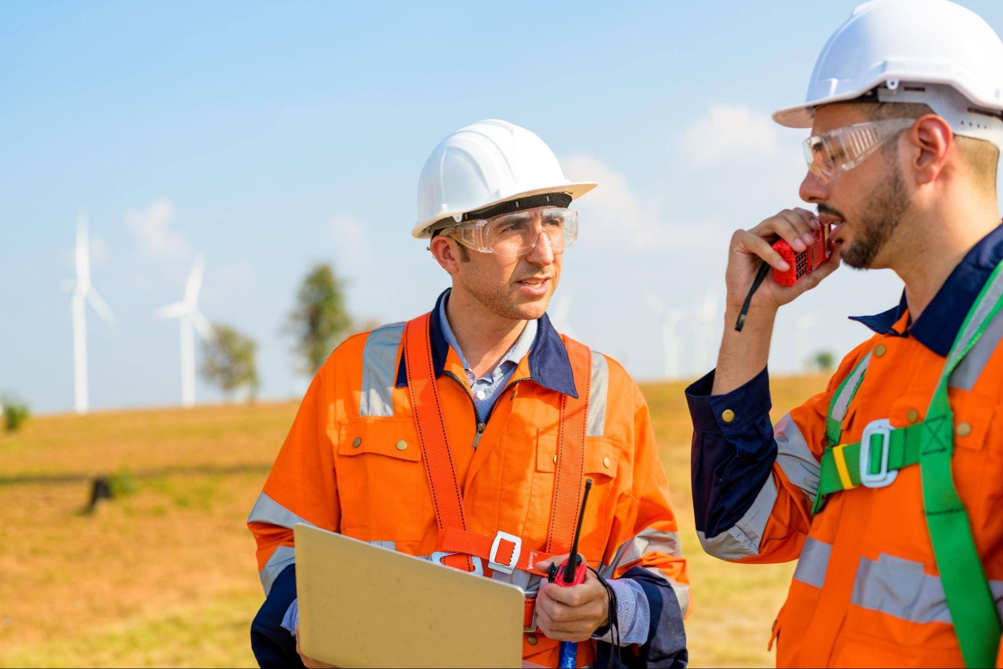 Two male field service technicians in full safety gear perform maintenance on a wind turbine in a renewable energy site.