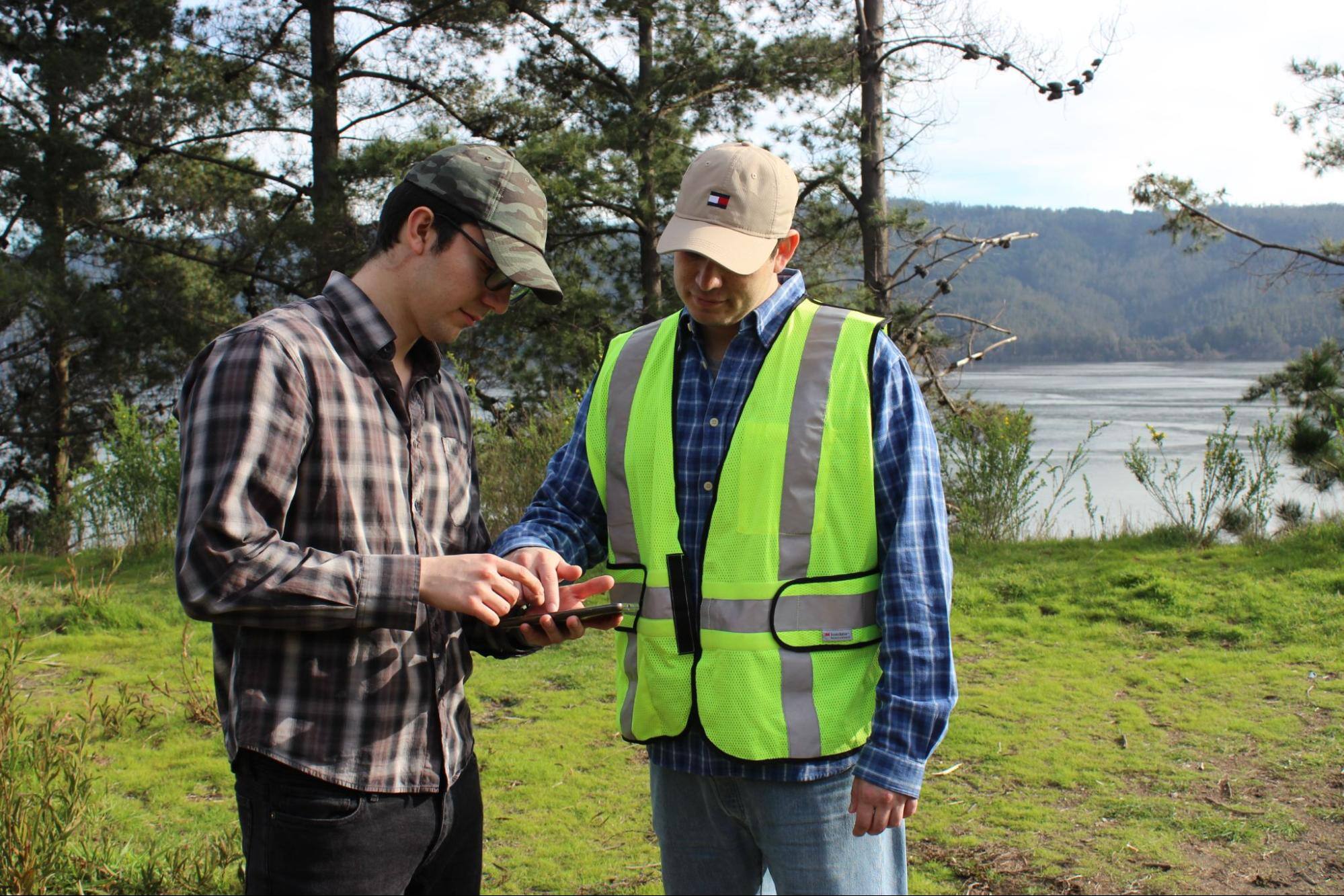 Two men looking at a cell phone with trees in the background. 