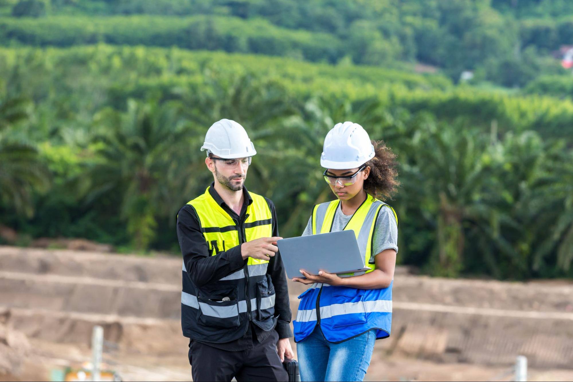 Two workers in hard hats looking at a computer outside.