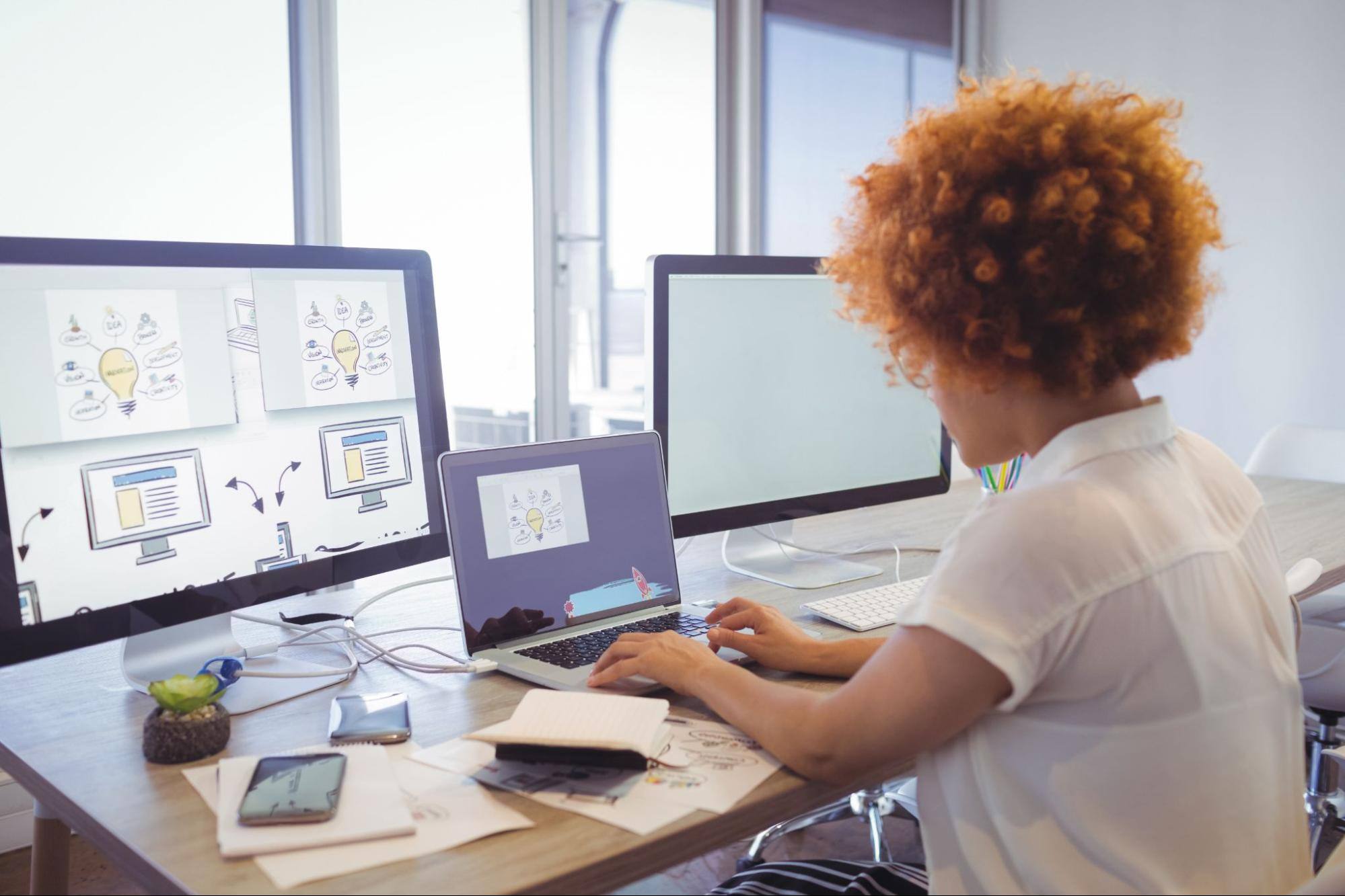 A woman at a desk using a laptop with two additional computer screens in front of her.