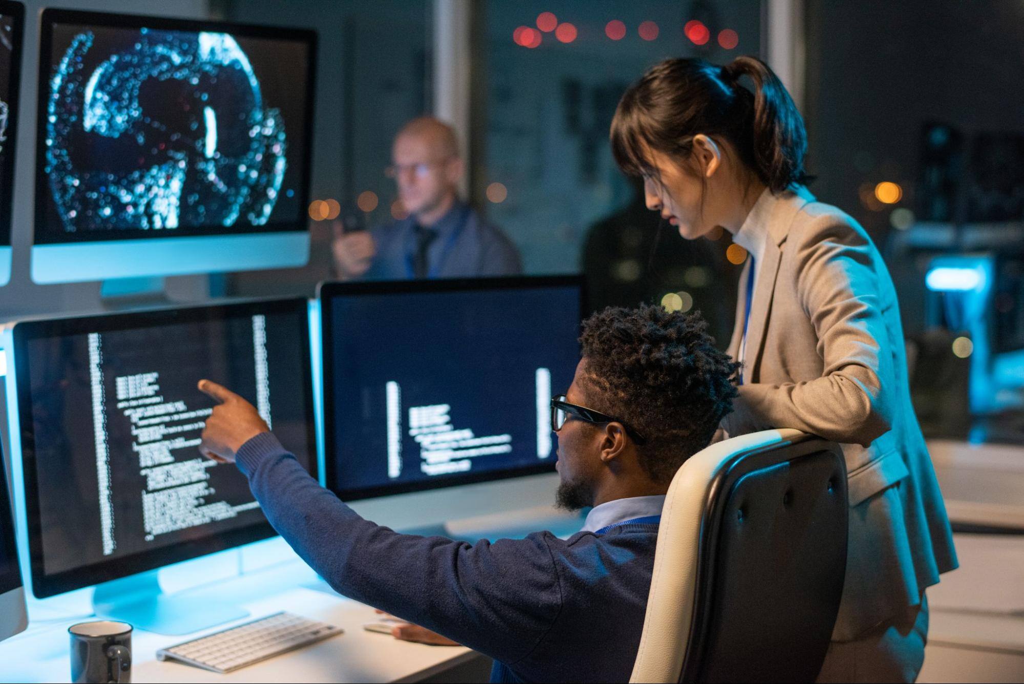 A man sitting in a chair and a woman standing in an office are looking at coded data on a computer screen.