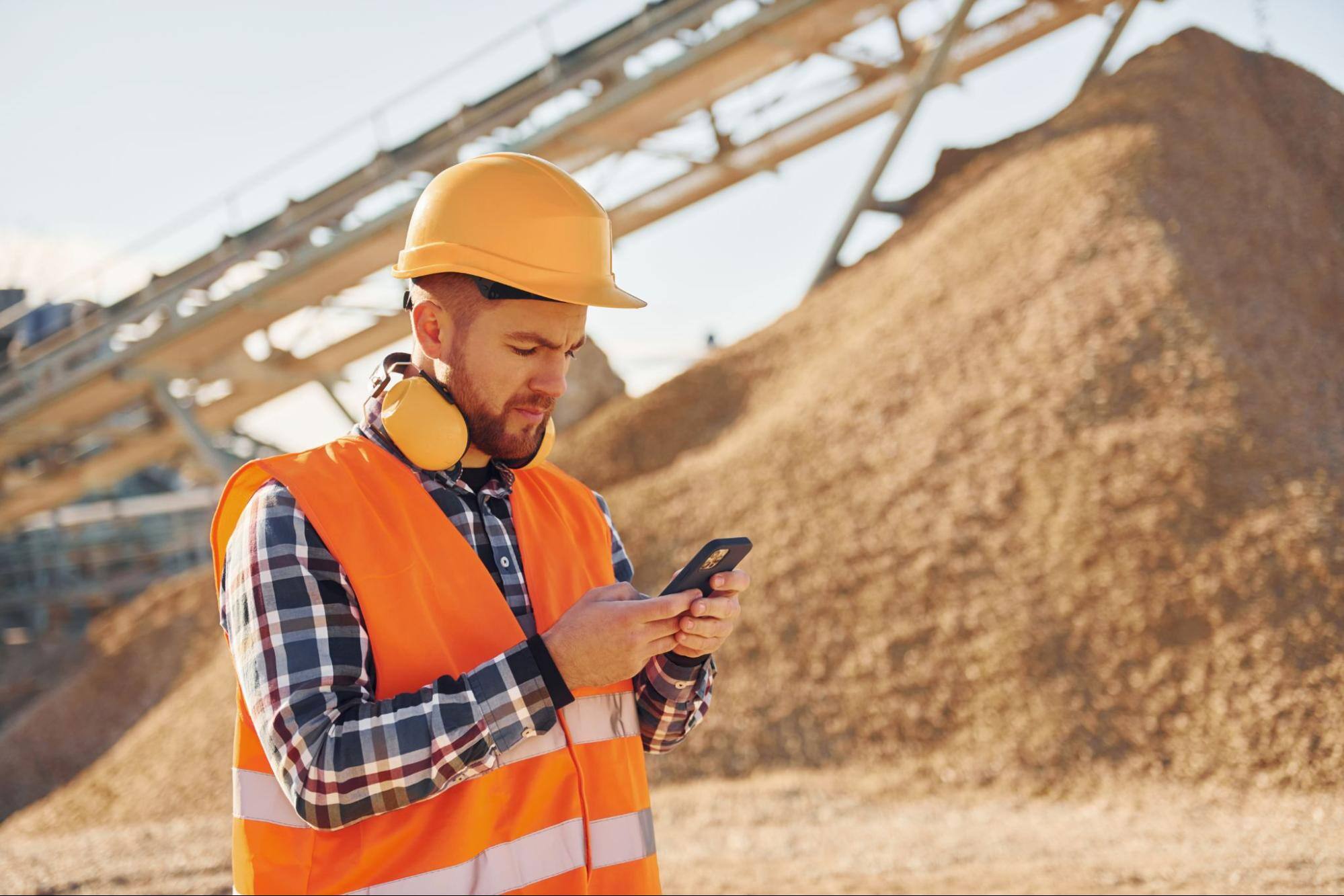 A construction worker in uniform, with yellow headphones around his neck, reports his findings to the primary office site using his mobile phone at a quarry site.