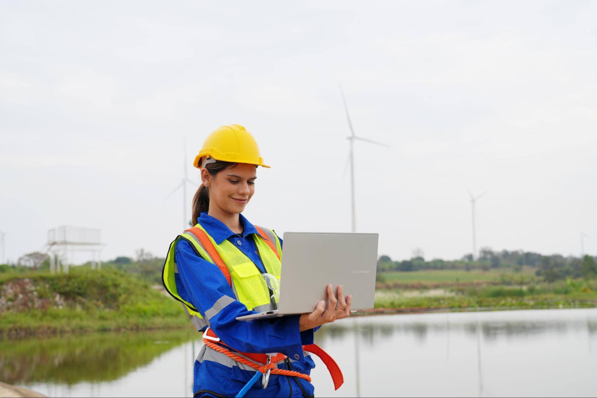 Female construction worker with a laptop outside next to a body of water. 