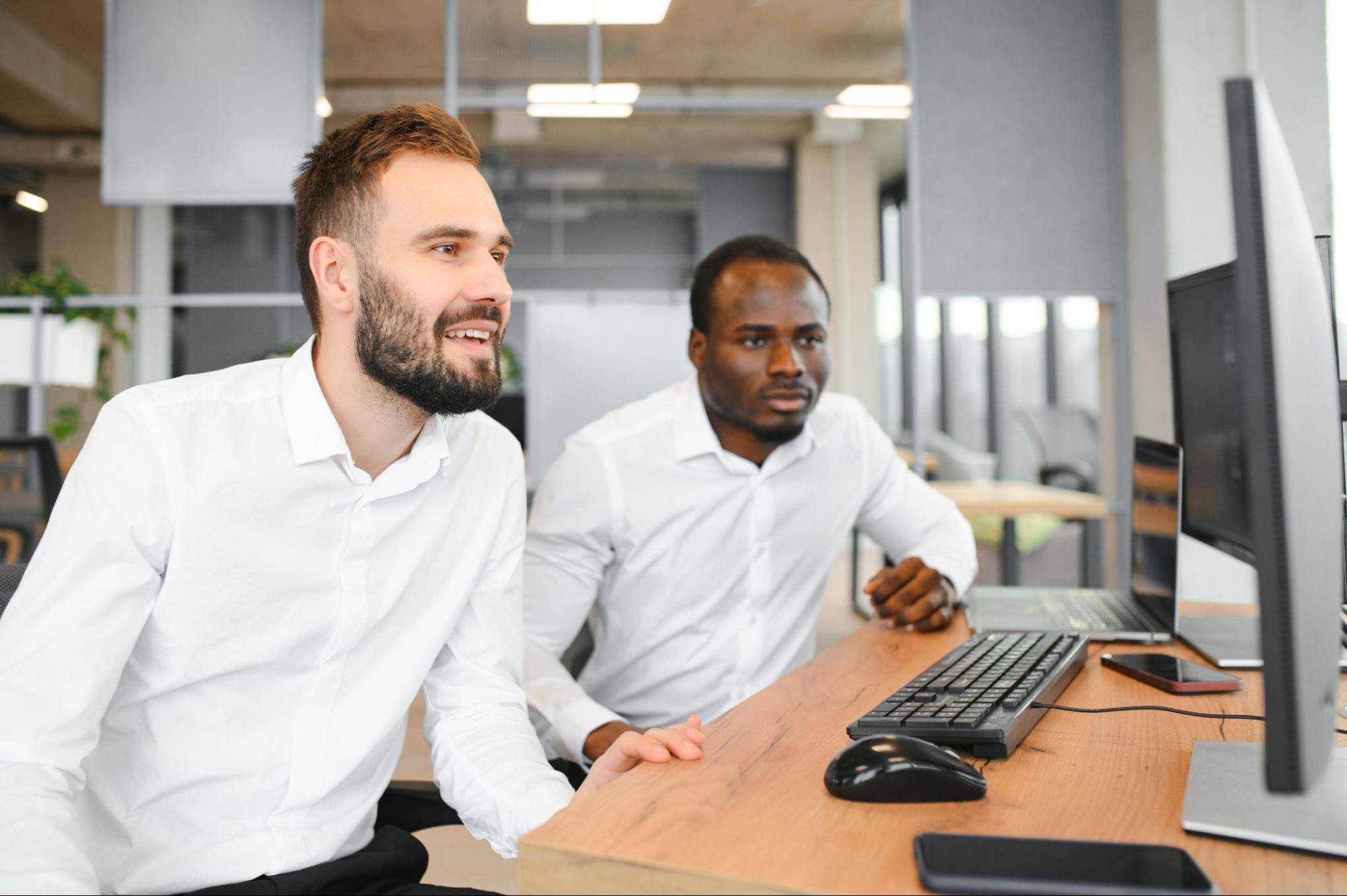 Two diverse colleagues, both traders, smile while sitting in front of their computers, pleased with the efficiency and benefits of automation tools.