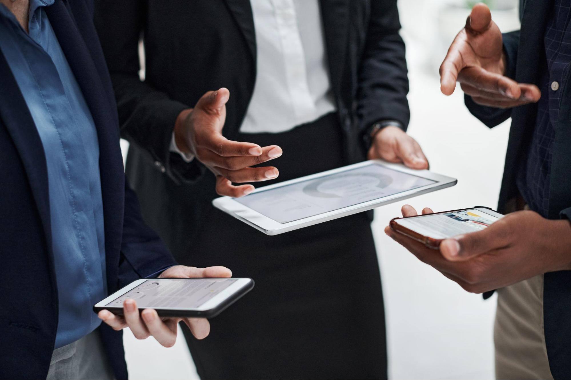 Cropped view of a woman’s hand holding a phone, a man holding a smart tablet in one hand and gesturing with the other, while another man has his phone and points his finger at the woman holding her phone.