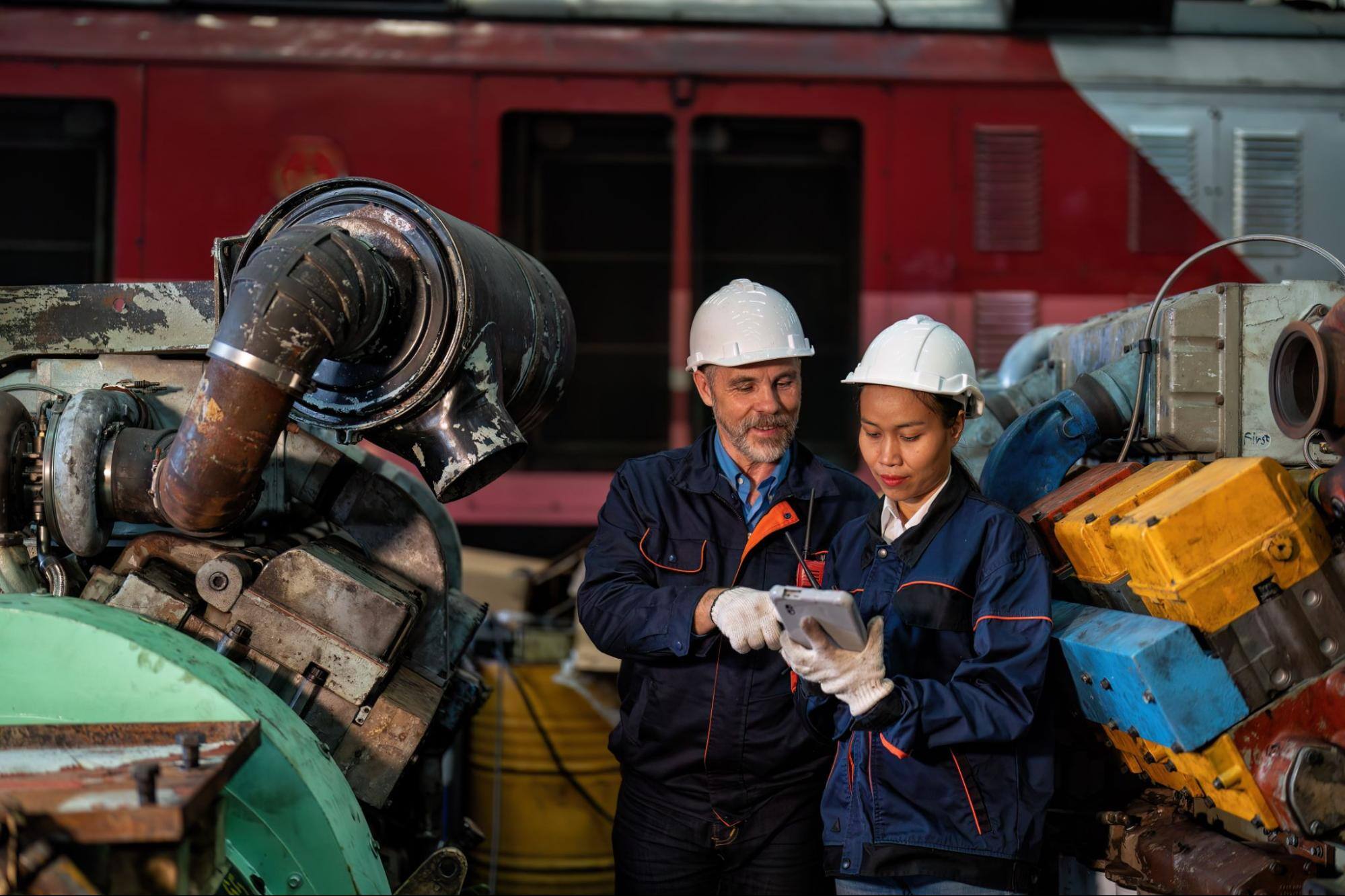 Two construction workers holding a device with machinery in the background. 