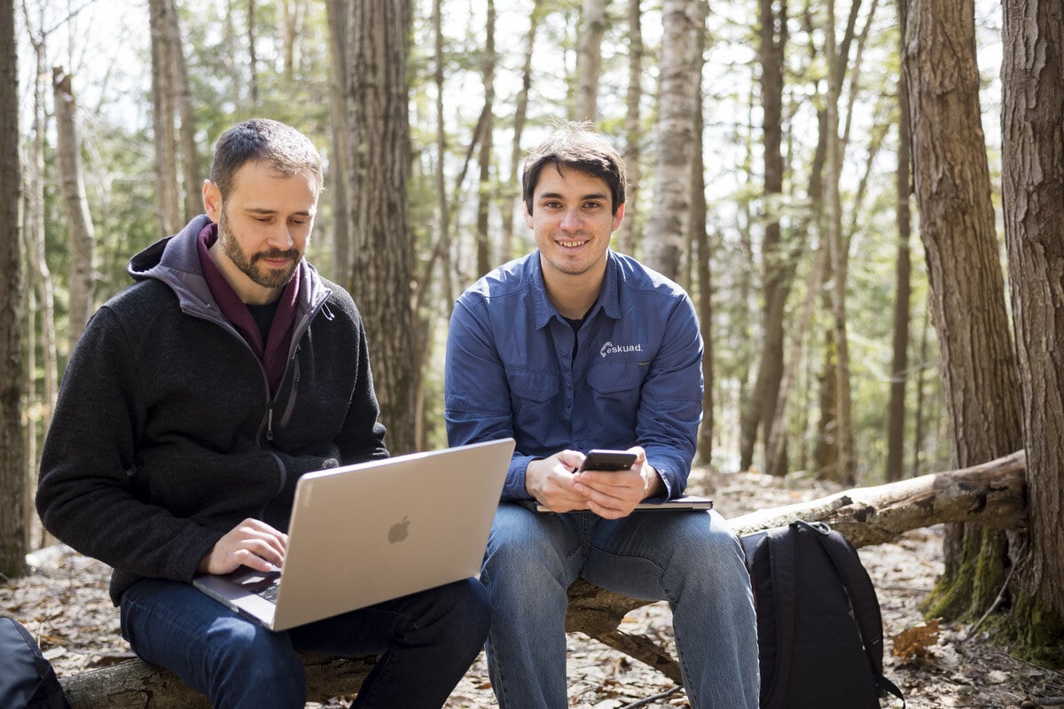 Two men outside, one with a computer and the other holding a cell phone. 