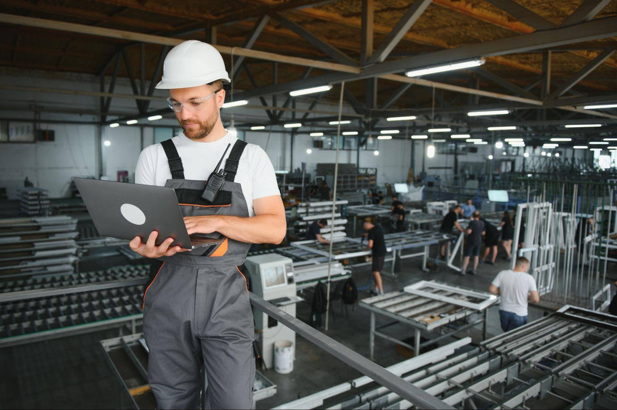 A smiling male industrial worker inputs data on his laptop in a factory, with steel forms and fellow workers visible in the background, highlighting automation in manufacturing.