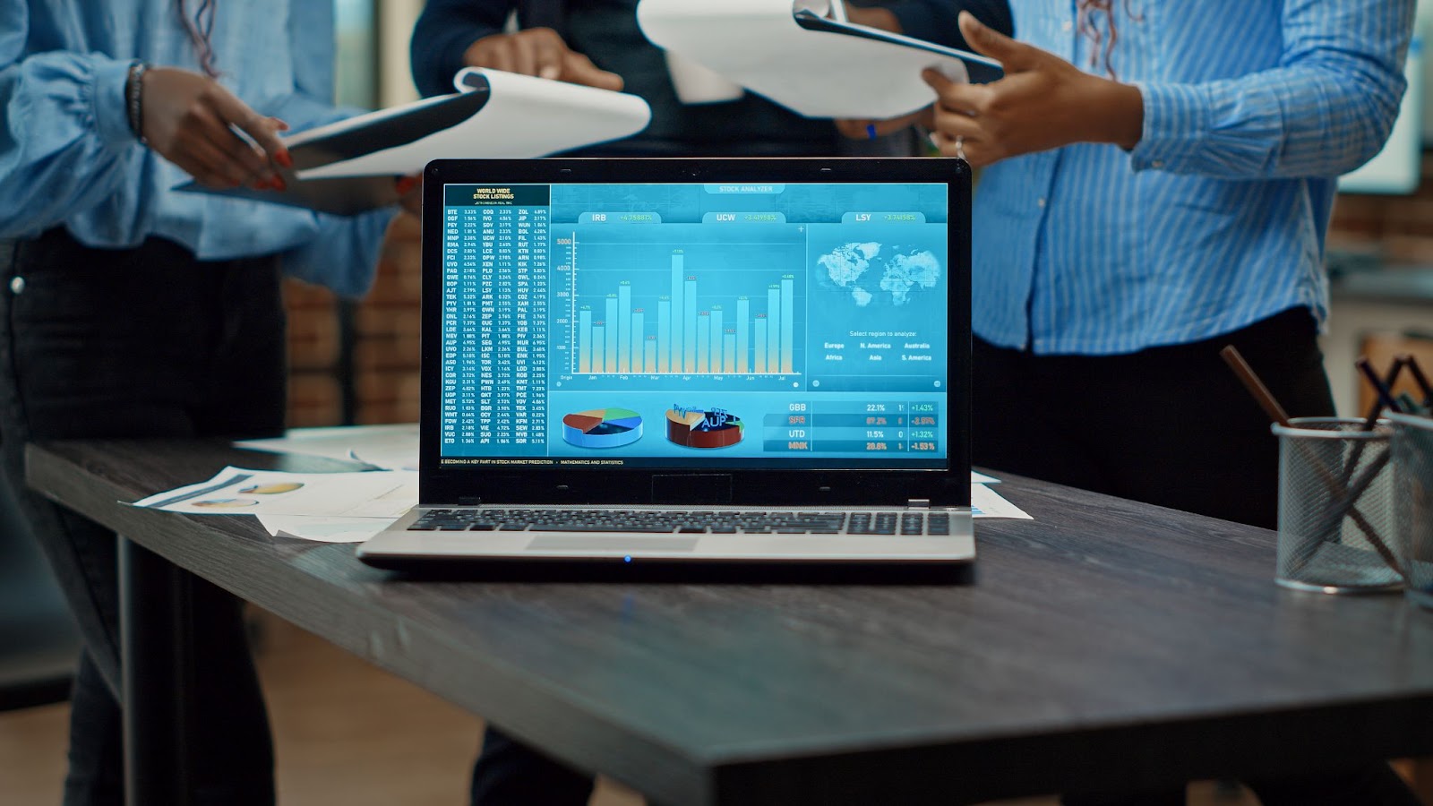  A close-up of a laptop displays online analytics data while three staff members stand behind it holding clipboards with raw information; their faces are not shown.